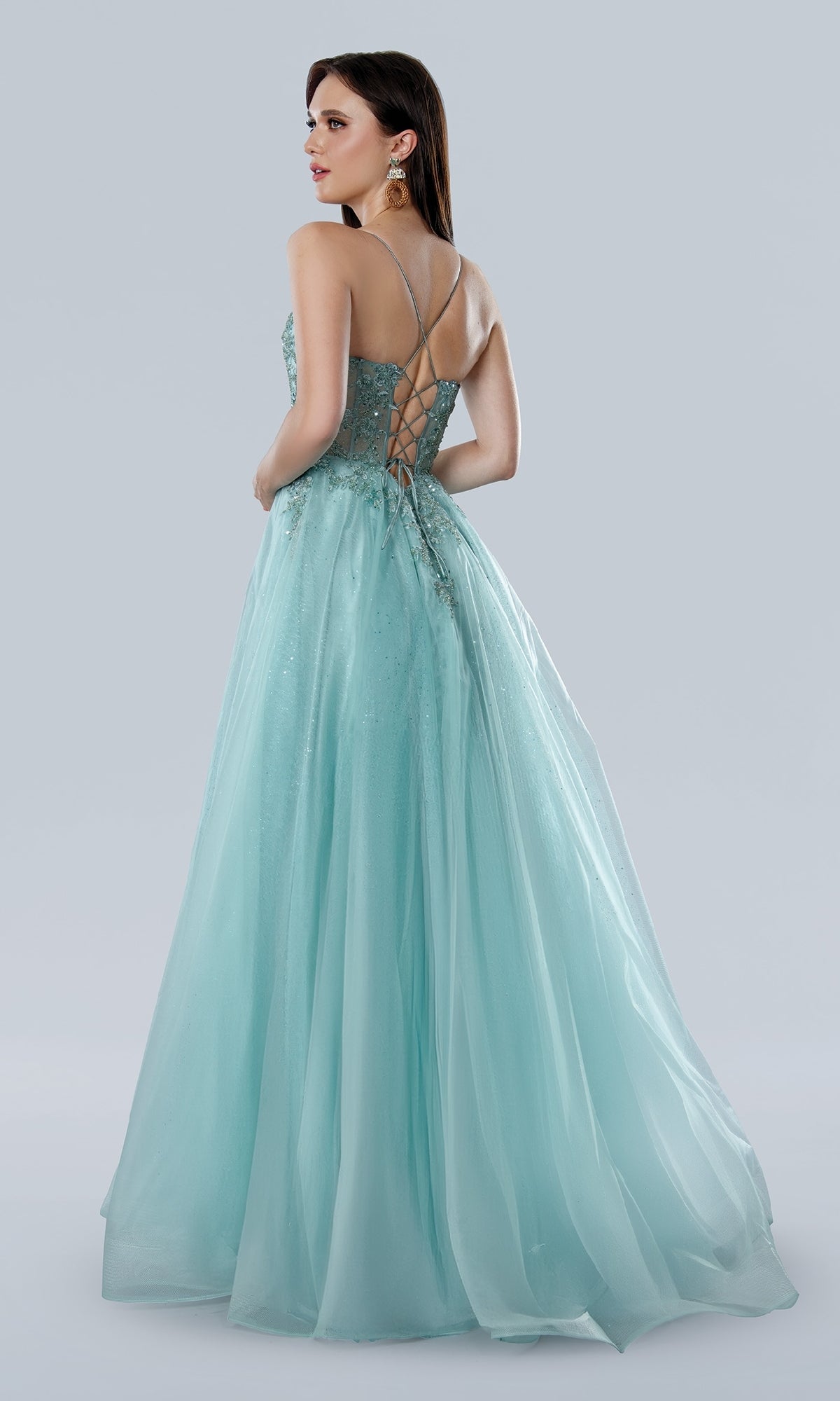 A woman stands on a plain light background wearing the Stella Couture 25140 Glitter Prom Ball Gown—a light blue, floor-length dress with spaghetti straps, V-neck corset bodice, and sequin lace embroidery.